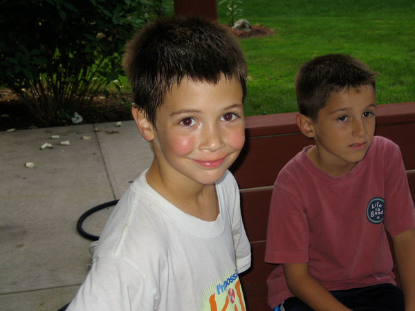 A young boy, with short hair and rosy cheeks, smiling up at the camera, another young boy sitting on a bench off to the side.