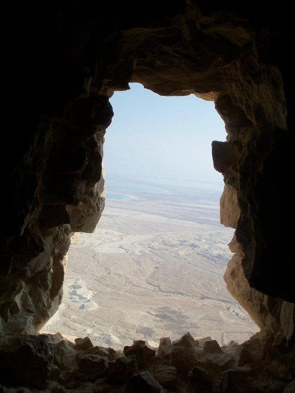 Inside a moutainous cave, looking through a small opening with a high view on the ground and water off in the distance.