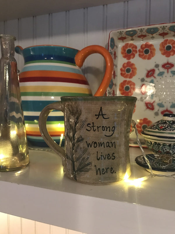 A mug alongside some other dishes that reads, "A storng woman lives here".
