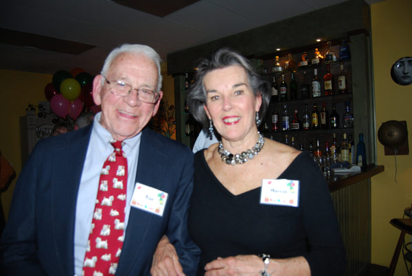An older man and woman arm in arm, wearing name tags, smiling, with balloons in the background.