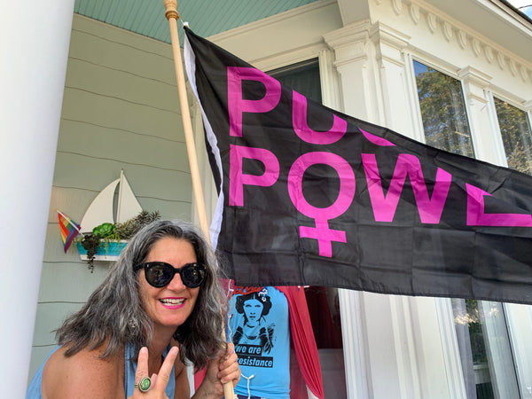 A woman smiling, giving the peace sign, holding a flag that reads "Pussy Power".
