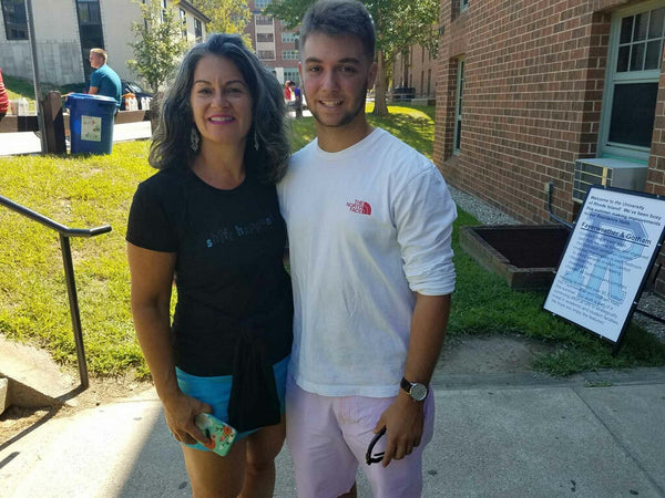 Woman in shorts and a t-shirt, volumous hair, smiling, her and her sons arm wrapped around each other, him smiling. A college poster on the ground behind them.