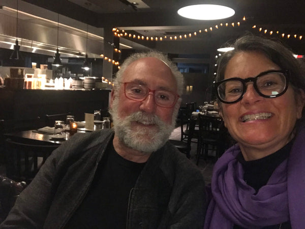 A woman in a colorful scarf and bold glasses, smiling next to an older man in glasses with a beard.