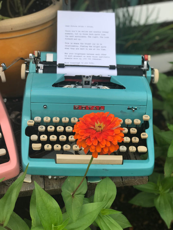 Outdoors, a typewriter with a bright flower growing in front of it.