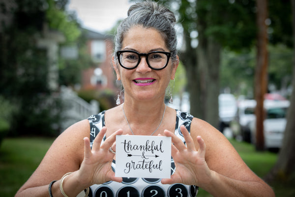 Woman with bold glasses, hair pulled back, smiling, holding a card that reads "thankful & grateful".